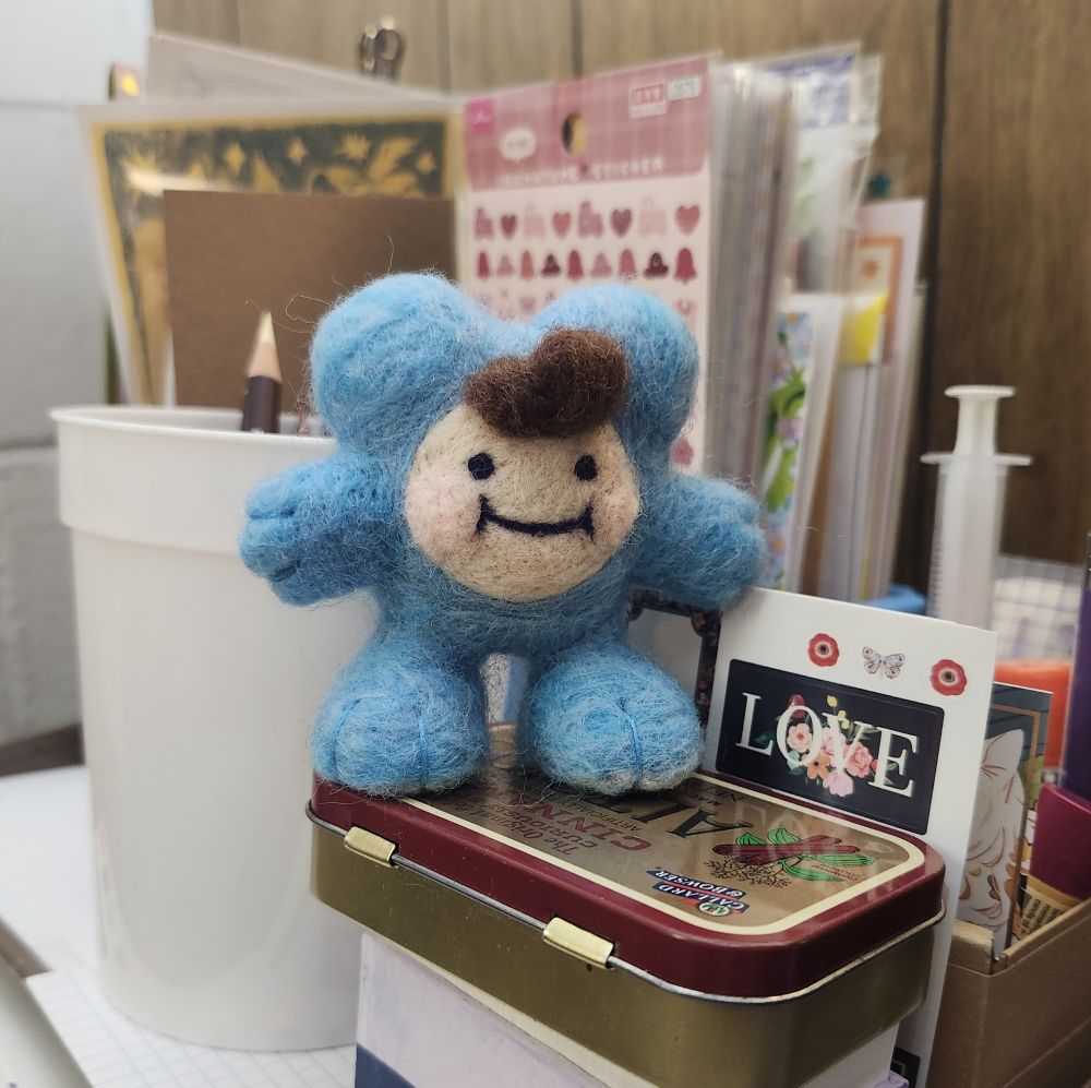 A small needle felted doll of CammyBoiG, who is a small round critter in a blue bear costume. He is standing on an Altoids tin.