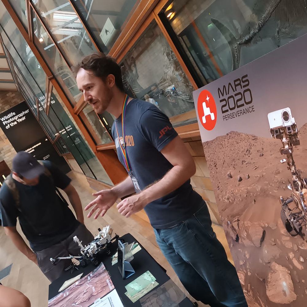Photo of me, a white man with brown hair wearing a blue “Mars 2020” t-shirt and jeans, standing behind a trestle table covered in images, maps, and a Lego model of a Mars rover. Next to me is a large poster board showing a selfie taken by the Perseverance rover, and behind me is a wall covered in ichthyosaur fossils in glass cases.