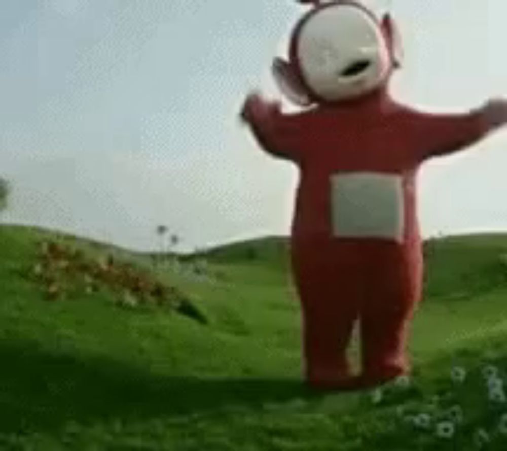 a red teletubbies teddy bear is standing in a grassy field with his arms outstretched .