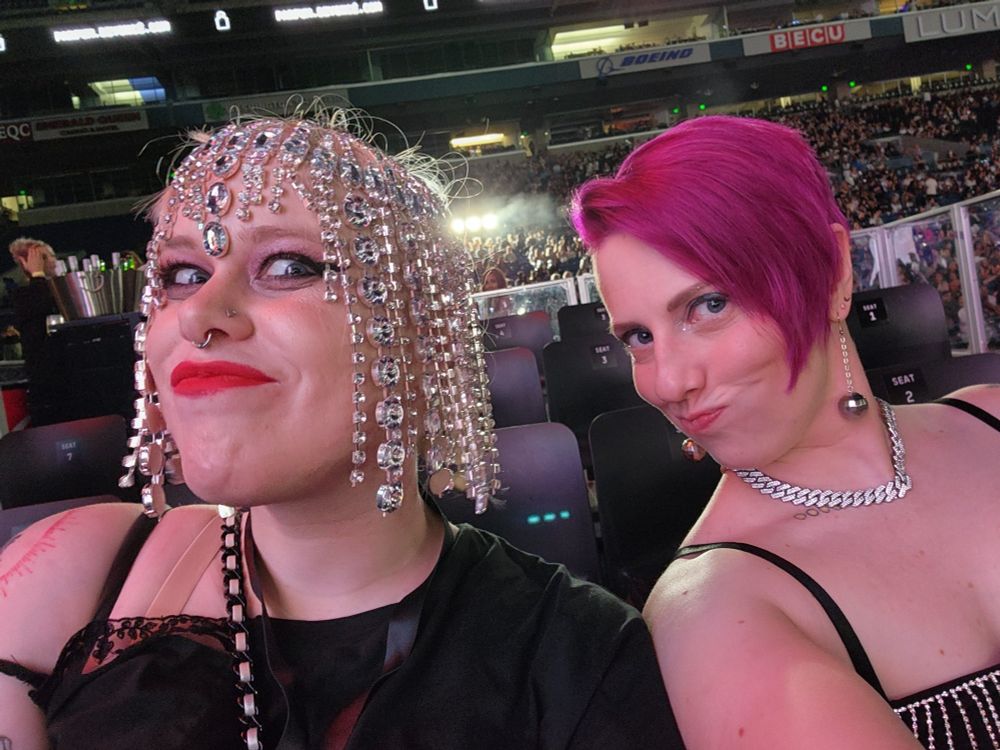 Two white milquetoast women in a selfie at a beyonce concert. One is wear a rhinestone headdress with glam makeup and the other has fushia hair in a 90s faux goth cut. Both look like absolute goblins as they contort their faces