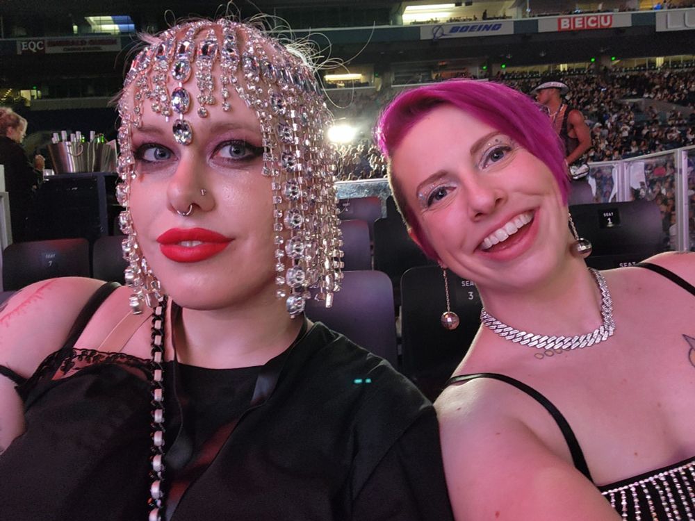 Two white milquetoast women in a selfie at a beyonce concert. One is wear a rhinestone headdress with glam makeup and the other has fushia hair in a 90s faux goth cut
