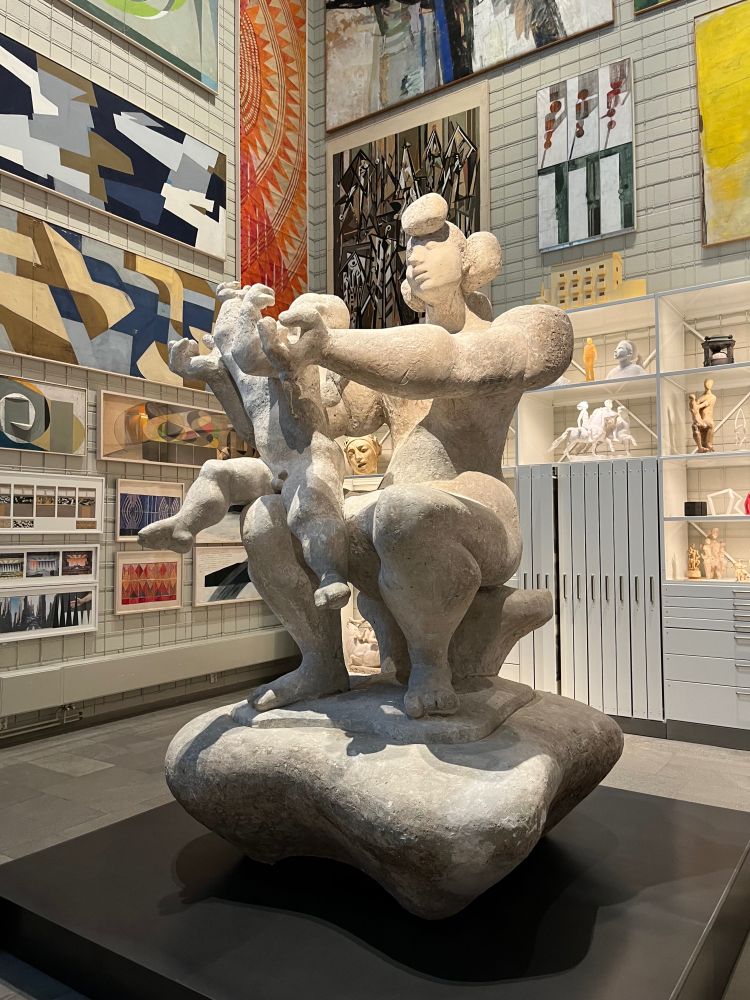 A figurative sculpture of mother and child intended for outdoor display, on a podium in a corner of an interior museum gallery. The walls behind are covered floor to ceiling in twentieth-century paintings of varied sizes.