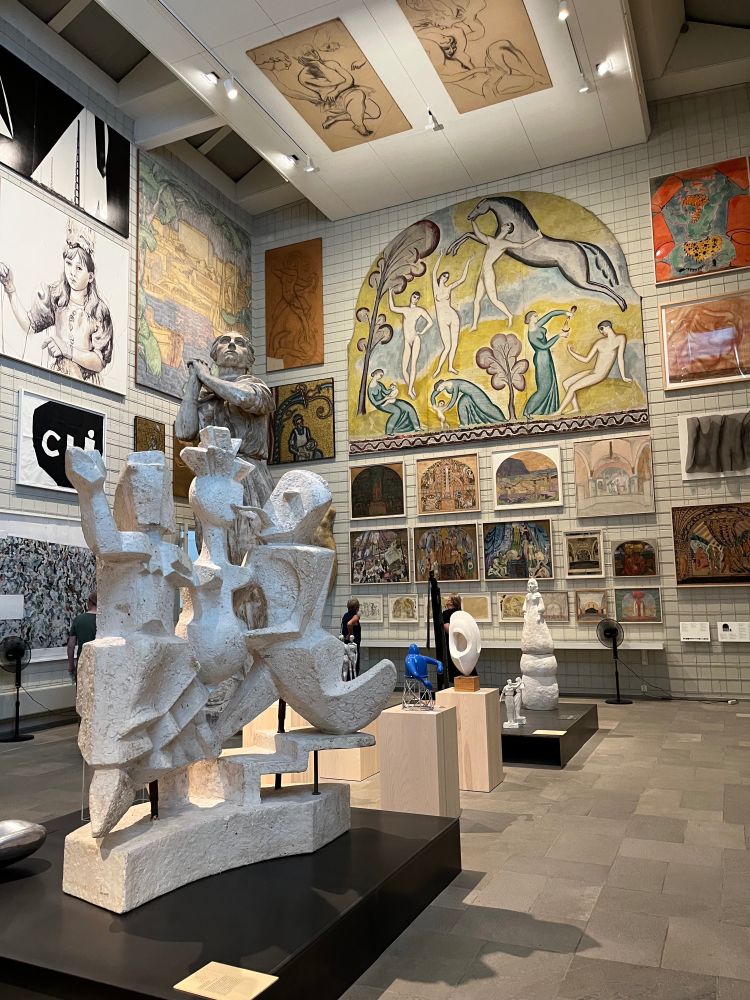 Photo of an interior corner of a large museum space with paintings on two walls and ceiling, and a series of twentieth-century sculptures in the middle of the room. 