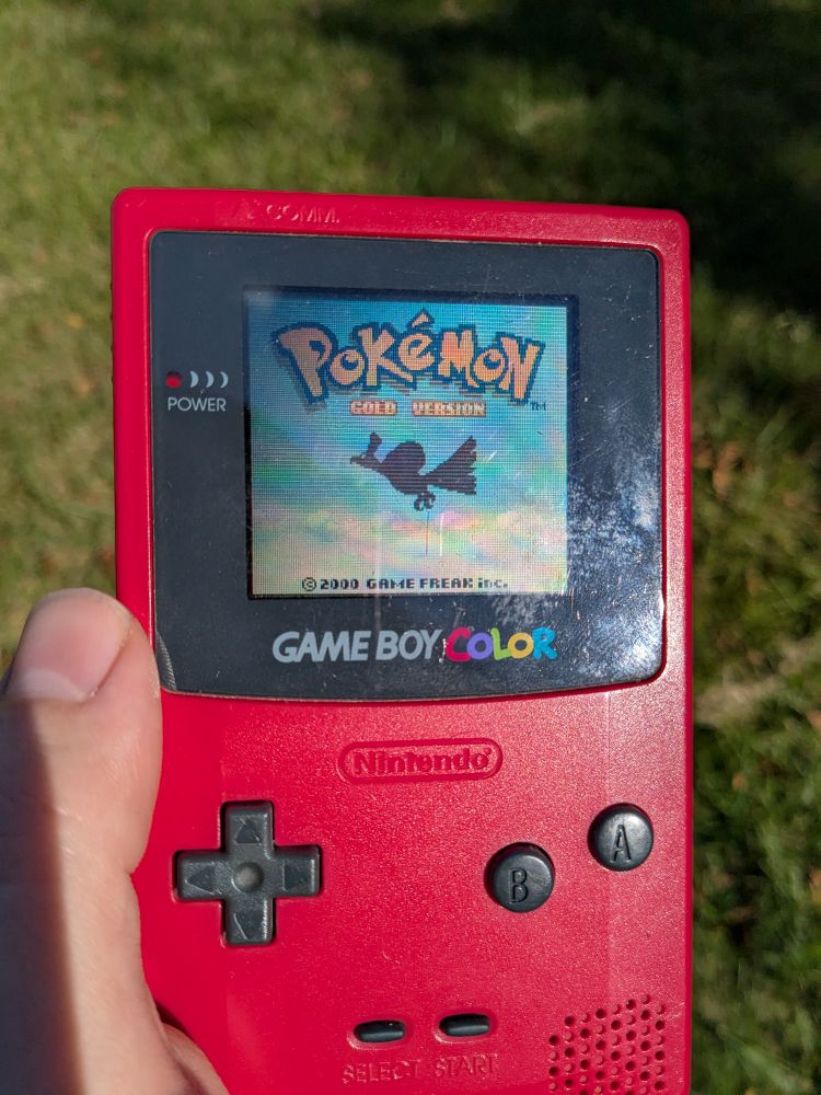 A red Game Boy Color showing the title screen of Pokémon Gold