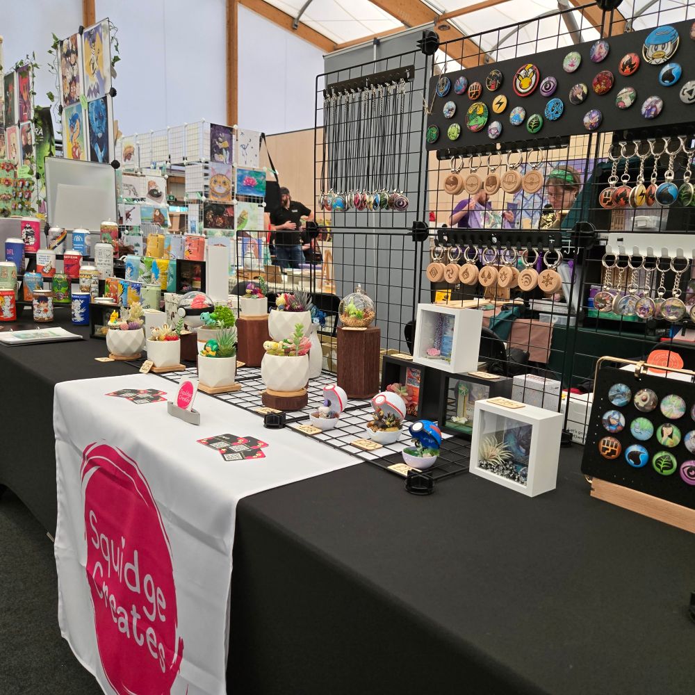 A photograph of the Squidge Creates trader stall at Canterbury Gaming Convention. The stall features Trinkets made from old trading cards, Pokemon terrariums and candles made from upcycled drinks cans