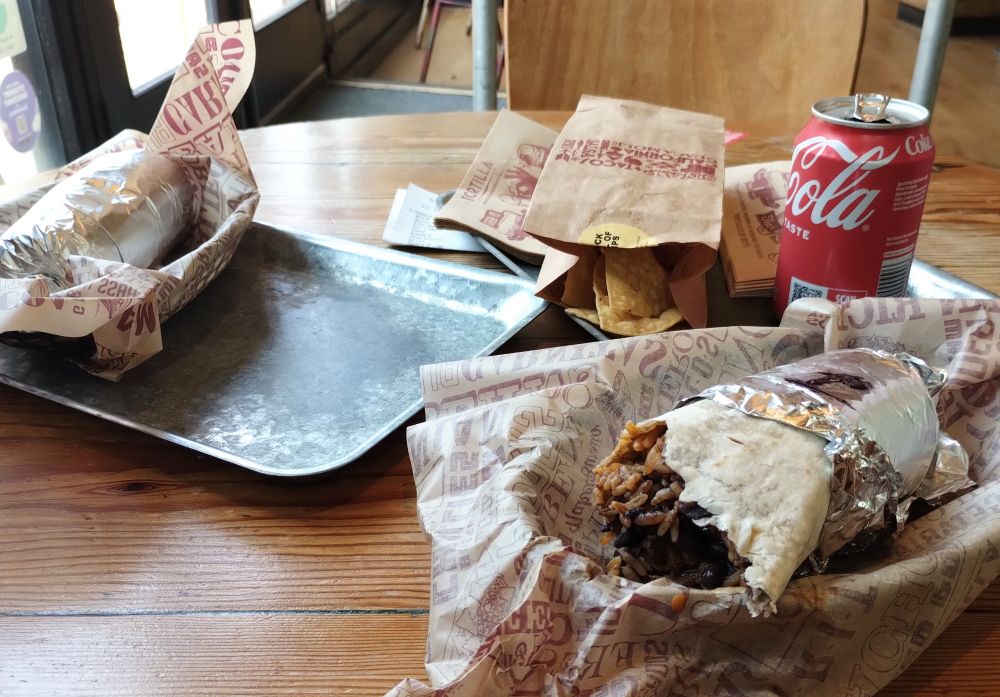 Two burritos on a wooden table - one started, the other still wrapped - plus a bag of chips and a can of coke.