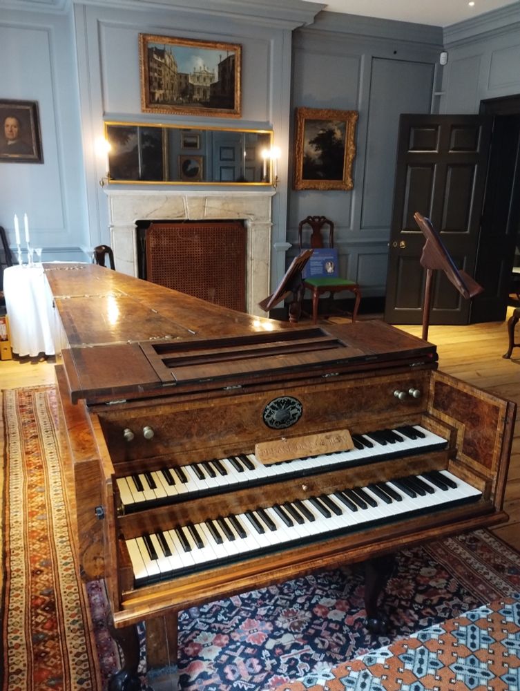 A large double harpsichord (with keyboards on two tiers) in a large formal room decorated as it would have been in the 1700s. Rugs on the floor, pictures on the pale grey panelled walls, and a white stone fireplace. A few chairs and music stands ready for rehearsals.