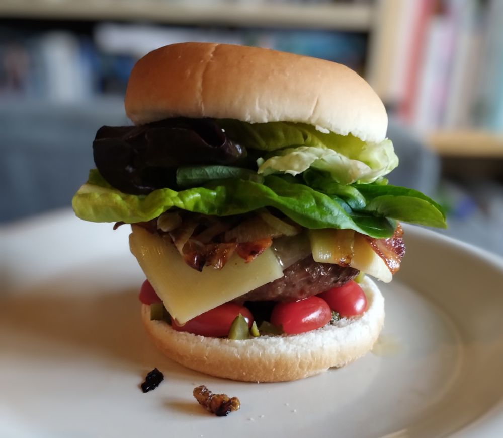 A beefburger in a white bun, stacked high, with halved cherry tomatoes, chopped cornichons, fried onions, bacon, cheddar and lettuce.