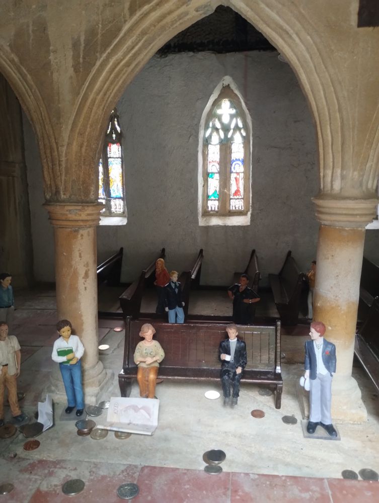 View into the window of a model church, with model people sitting in pews surrounded by gigantic (real) money which has been dropped through a slot higher up. Behind them, light comes through miniature stained glass windows.