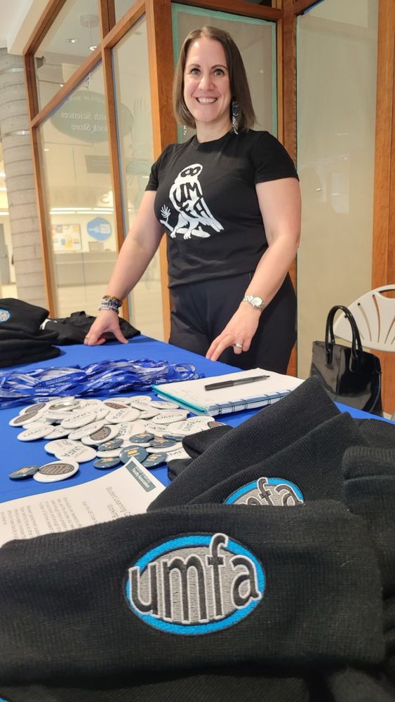 UMFA past-president Orvie Dingwall stands at a table with UMFA pins and flyers. She is wearing an UMFA tshirt.