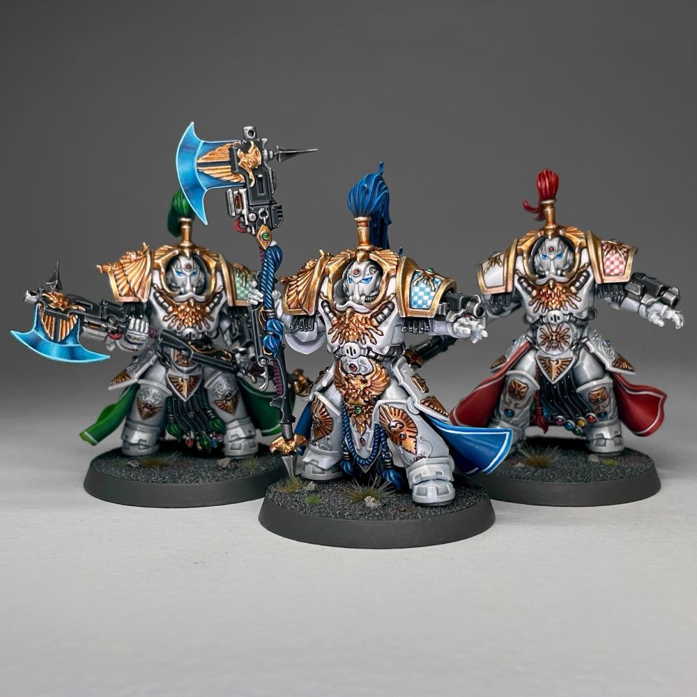 An Allarus terminator squad comprised of three Custodians, each in white and gold armour with different coloured robes. The model in the foreground has blue robes and plume while the others are red and green.