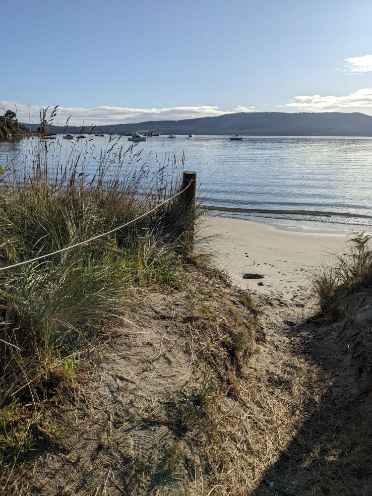 Sandy path to beach, calm water blue sky, boats in background