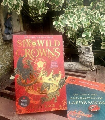 A paperback copy of "Six Wild Crowns" by Holly Race is displayed outdoors on a rustic wooden bench, with a leafy variegated plant in the background. The red and gold cover features a dragon encircling a crown of six queens. Next to the book is a companion booklet titled "On the Care and Keeping of Lapdragons." Decorative elements include a small owl ornament and a fairy figurine on the wooden wall.