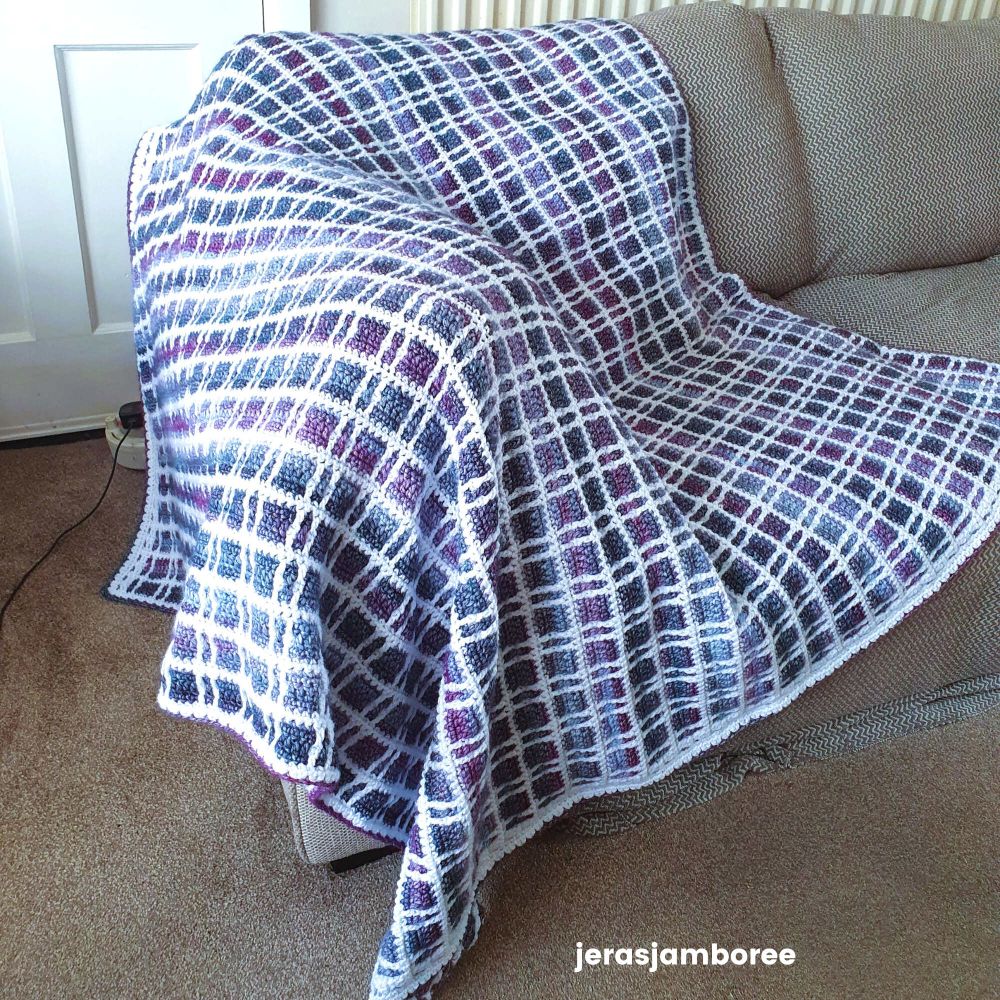 Fireside Plaid Mosaic Crochet Blanket displayed on a sofa, showing its purple, blue, and white tartan design