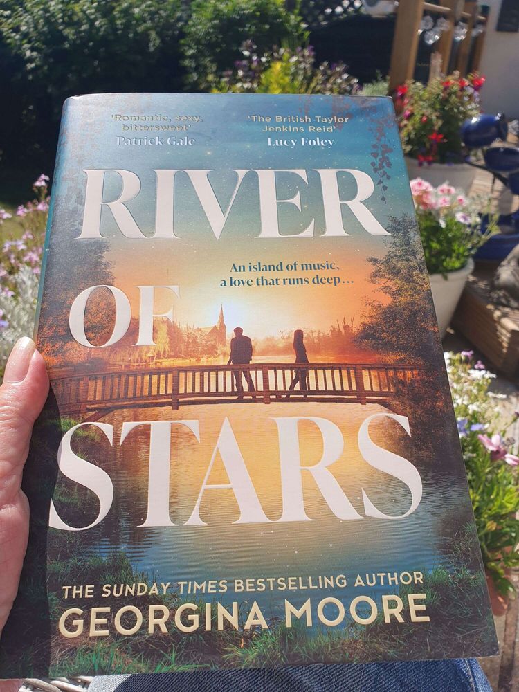 A person holds a hardback copy of River of Stars by Georgina Moore in bright sunlight. The book’s cover shows two figures on a bridge over water at sunset, surrounded by trees. Flowers and garden furniture are visible in the sunny background.