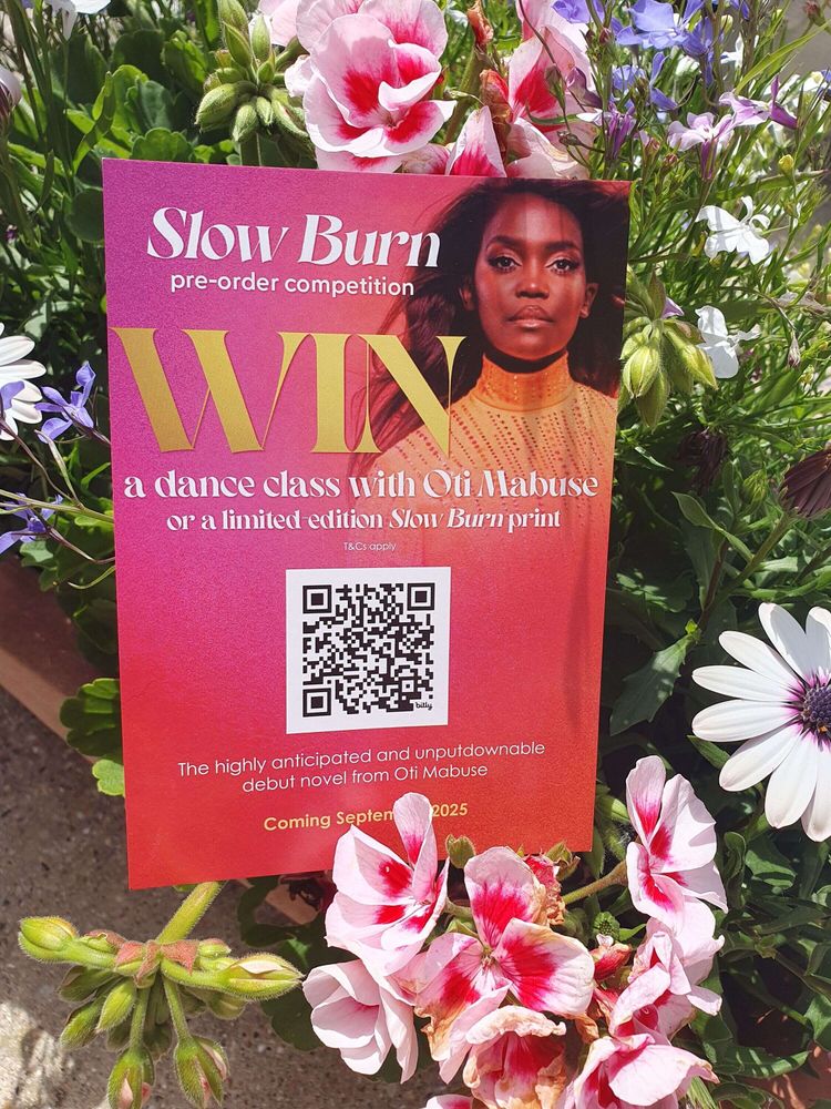 A vibrant promo card for the Slow Burn pre-order competition is nestled among pink and white flowers. The card features a bold photo of Oti Mabuse and text announcing a chance to win a dance class with her or a limited edition print. A QR code is centered below the details, alongside the release date: “Coming September 2025.”