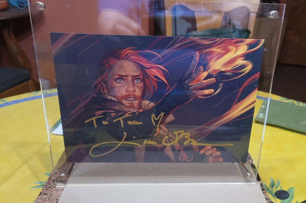 Photo of a print of a painting of Caleb Widogast slinging fire, signed in gold by Liam O'Brien and addressed to Tom with a heart, in a clear acrylic floating frame standing on a round table. That's 8 prepositions for those playing the home game.