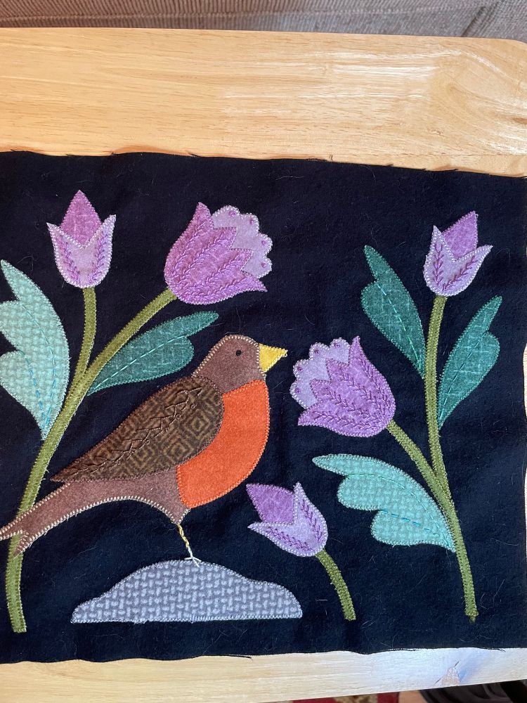 A machine appliqué quilted block showing a robin perched on a rock surrounded by purple and green tulips, set on a black background. The bird, flowers and leaves have decorate hand-stitching 