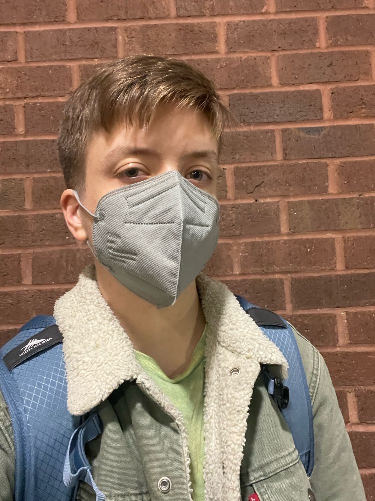 Eli as an extremely normal blond white boy in a gray KN95, green jacket with fleece collar, and blue backpack