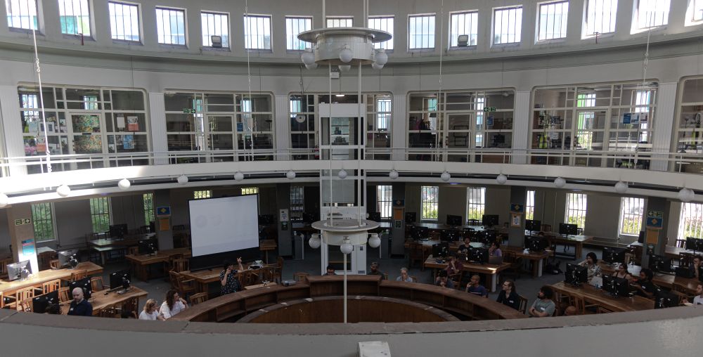 An image of the round reading room at the University of Glasgow.
The building is a circular room with art-deco architecture and details. People are sitting at computers on circular benches arranged concentrically, and a presenter is standing next to a screen.