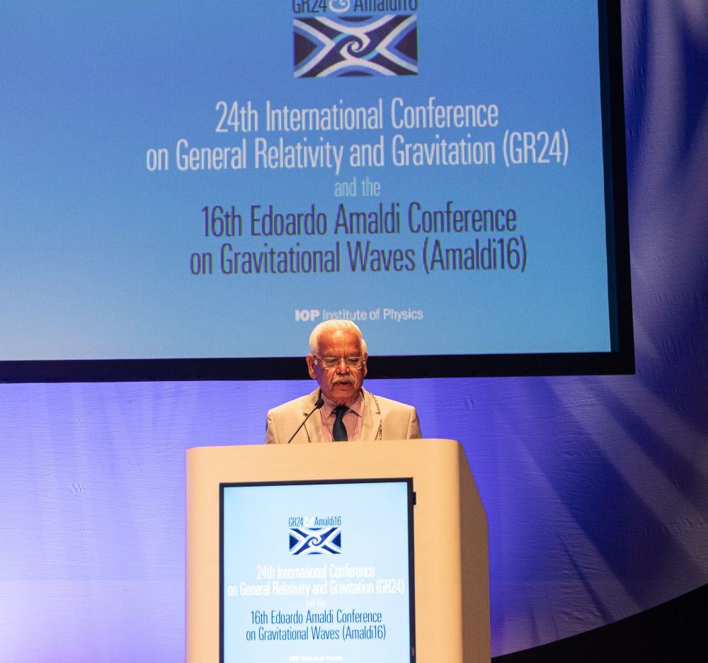 A photograph of Bailie Hanif Raja standing at a rostrum with the GR-Amaldi logo projected behind him.