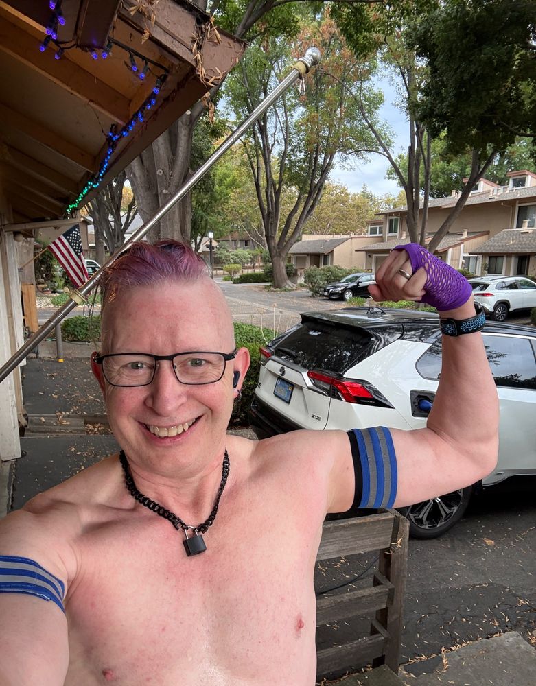 Selfie of a smiling shirtless man with purple hair, wearing blue reflective armbands and purple mesh gloves, flexing his left biceps. He has glasses, and a black padlocked chain collar around his neck. 