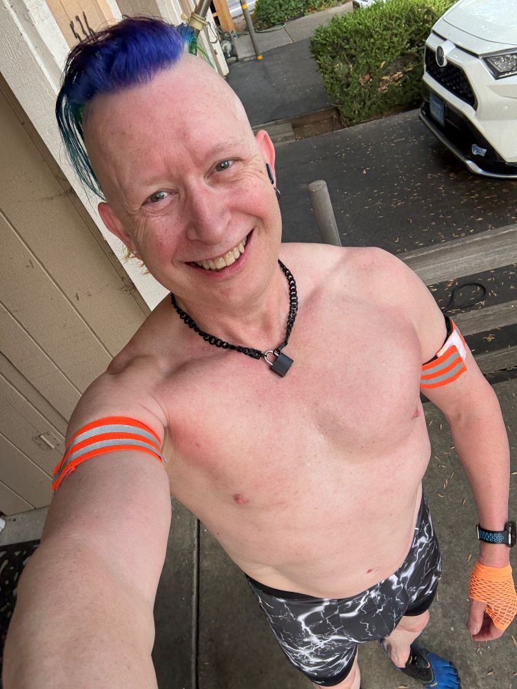 Selfie from above of shirtless Qubit grinning in compression shorts covered in white “lightning” on a black background