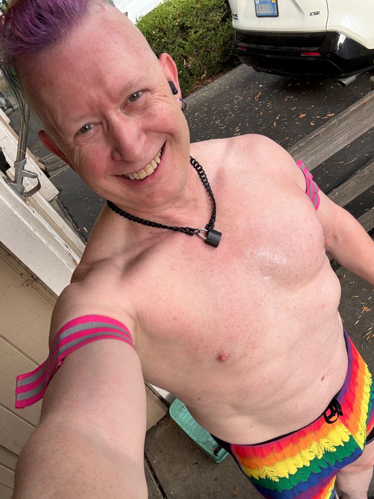Selfie from above of a smiling shirtless man wearing a black padlocked chain collar, pink visibility armbands and gloves, rainbow compression shorts and a tank top with a dancing rainbow astronaut on it. 