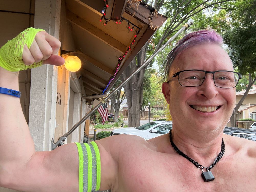 Closeup of barechested qubit smiling and flexing his right biceps. He’s wearing yellow hi-viz mitt and armband, and has a black padlocked chain collar around his neck 