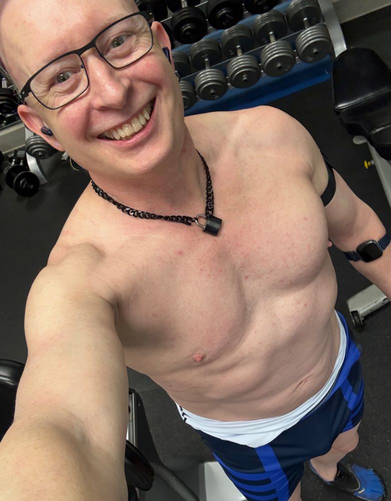 Photo from above of a smiling slender muscular man in a blue wrestling singlet rolled down to the waist in front of a rack of dumbbells 