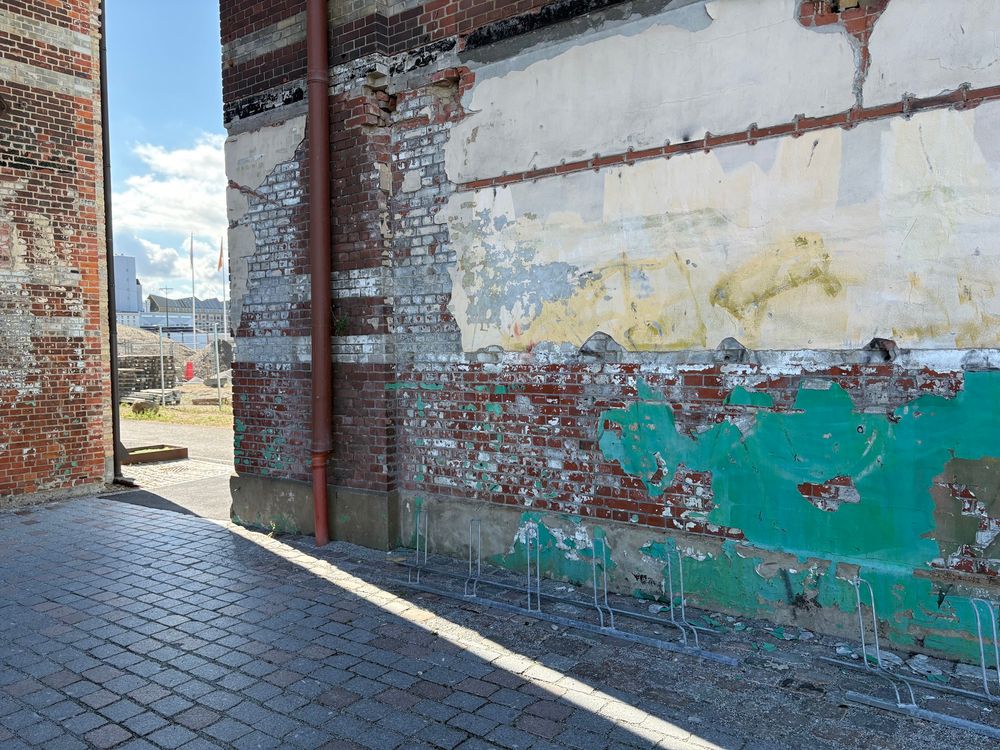 A corner of an old brick warehouse. The paint on the wall has the colours of a seascape: white-gold sky above, greenish sea below. 