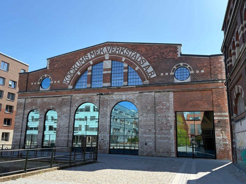 Old industrial building, with the name of the firm Kockums. It has high, arched windows: the glass reflects the modern buildings behind, and above those, the sky.
