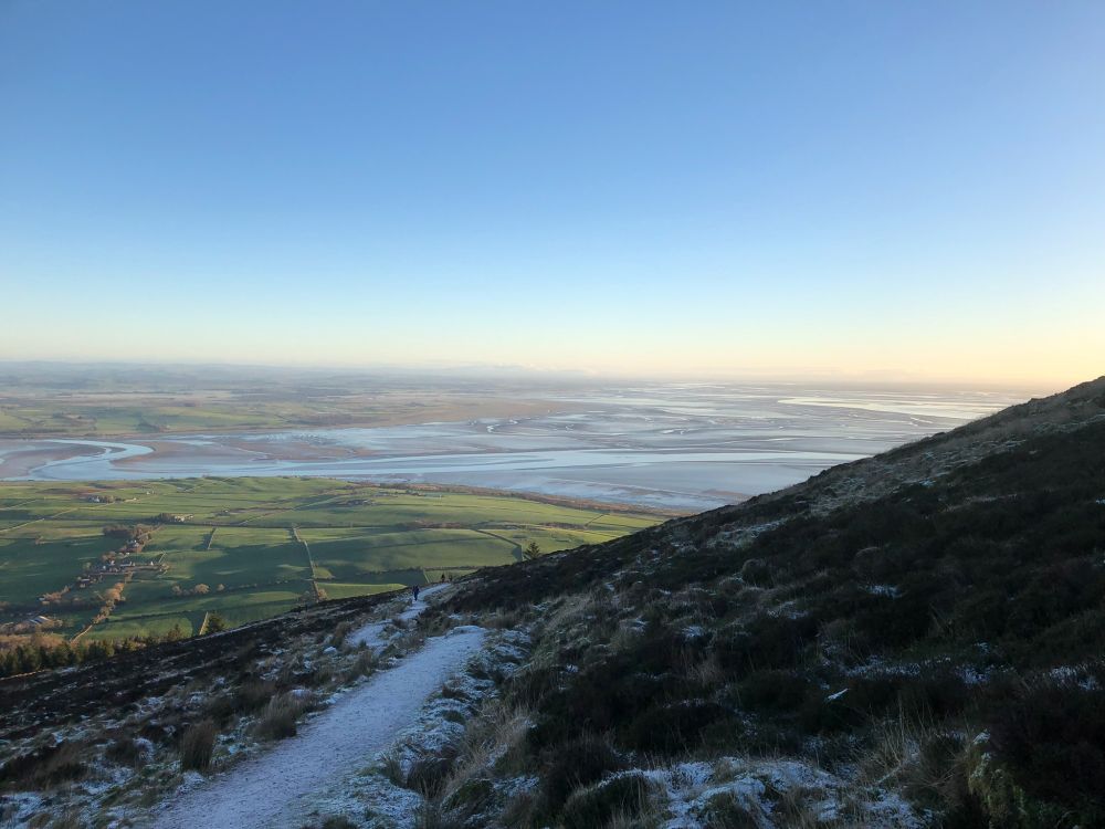 An icy path heading up the shaded side of Criffel, with the channels of the river Nith heading into the Solway Estuary in the background