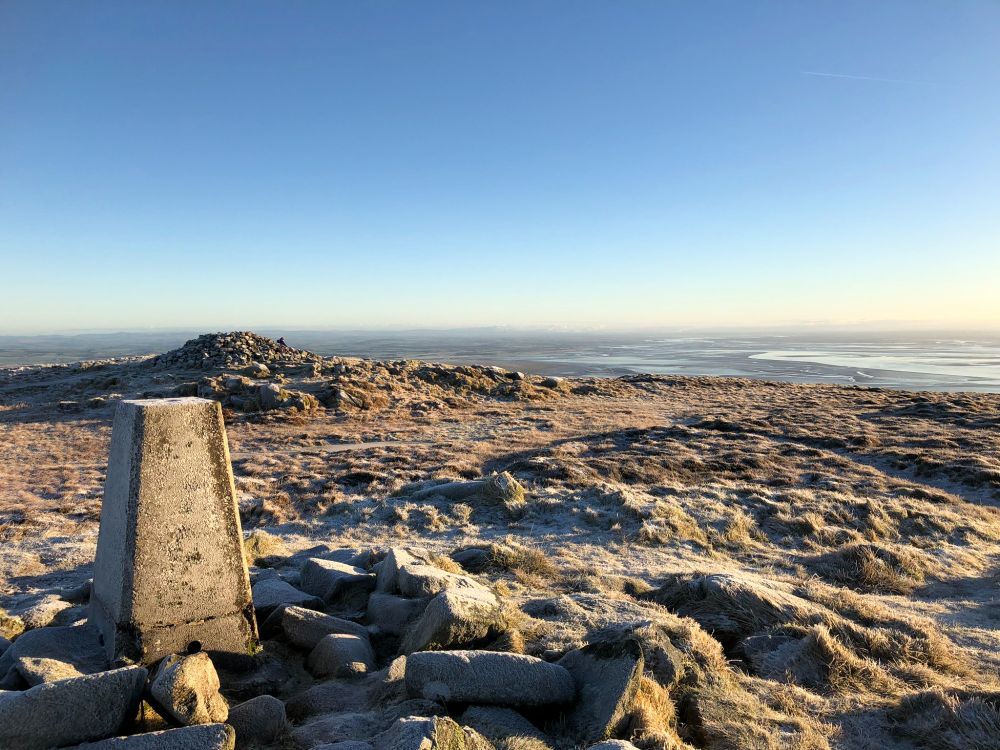The trig point of Criffel in the bottom left foreground, with the cairn behind it. A think layer of frost is on the ground, and the channels of the Solway Firth are visible in the background below a clear blue sky.