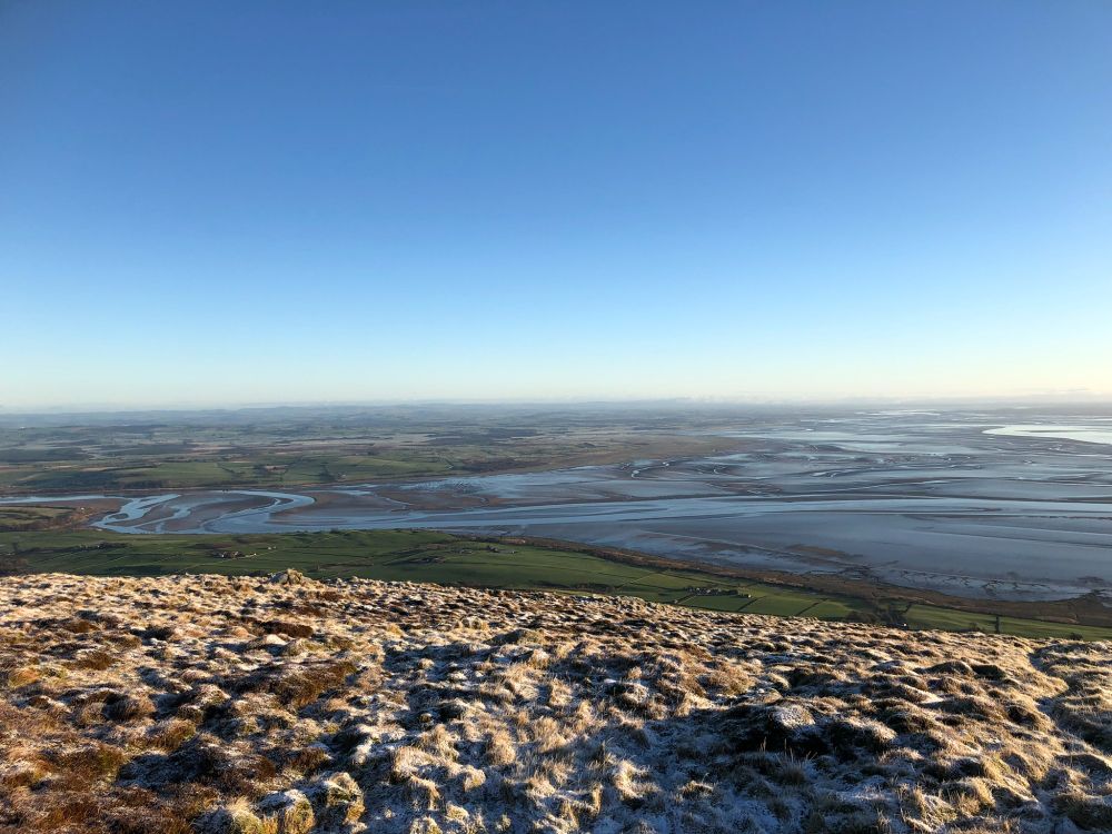 Viewed from the frosty front flank of the top of Criffel, the green fields of the short plain below contrast. The channels of the river Nith are visible in the Solway Estuary behind under a clear blue sky. 