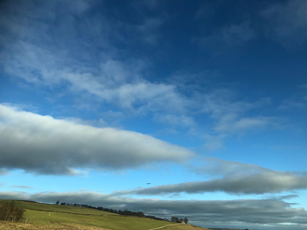 A military transporter plane performing manoeuvres over Caldbeck Common against a bright blue sky with large isolated grey clouds. 