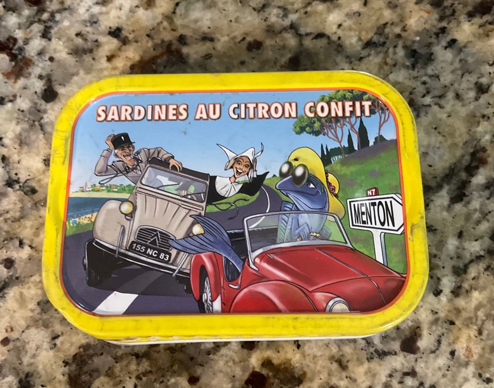A tin of sardines. The artwork shows a French policeman and nun driving a car, chasing after a fish in a jaunty hat and glasses, also driving a car 