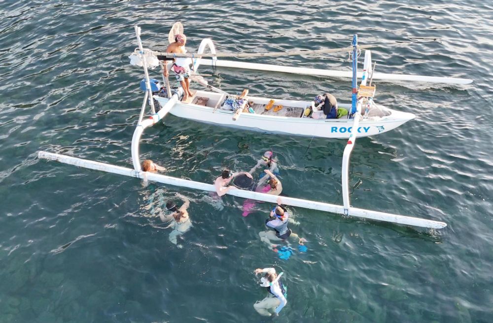 People in the water surrounding a fishing boat, Lee is on the boat drawing what they describe. 