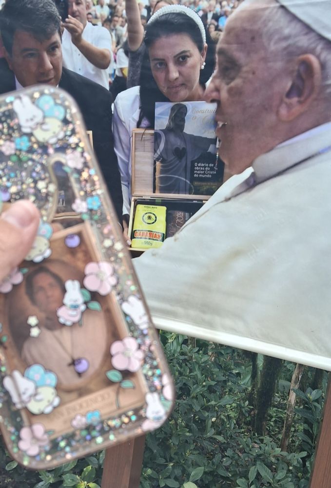 Uma capinha transparente cheia de glitter e desenhos de coelhinhos com a foto do personagem Vincent Benítez nela. Atrás, mais nítido, um poster com a foto do Papa Francisco.