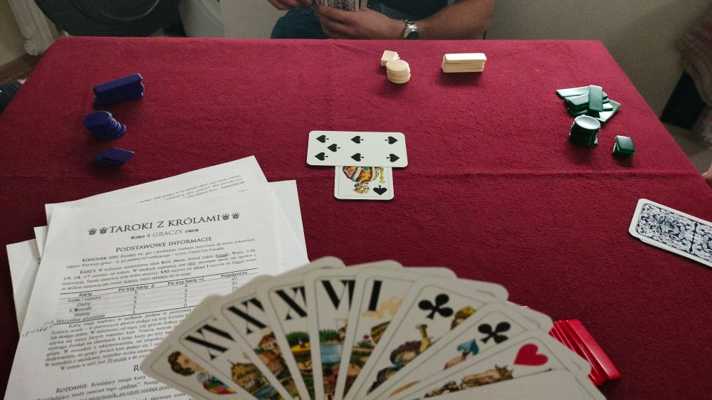 A first person view of a table with a burgundy cloth. Four people sit by it, playing cards, a tarot game, three of which are out of frame and the fourth is only partially visible. There are four sets of chips on the table in different shapes and colours. There is a bunch of paper with printed game rules in the bottom left corner. In the middle of the table there is an unfinished trick, lead with a king of spades, then followed by a 7 of spades. The player to the right has a captured trick on their side of the table. In the foreground there is a fanned hand of cards consisting of six tarocks and four plain suited cards.