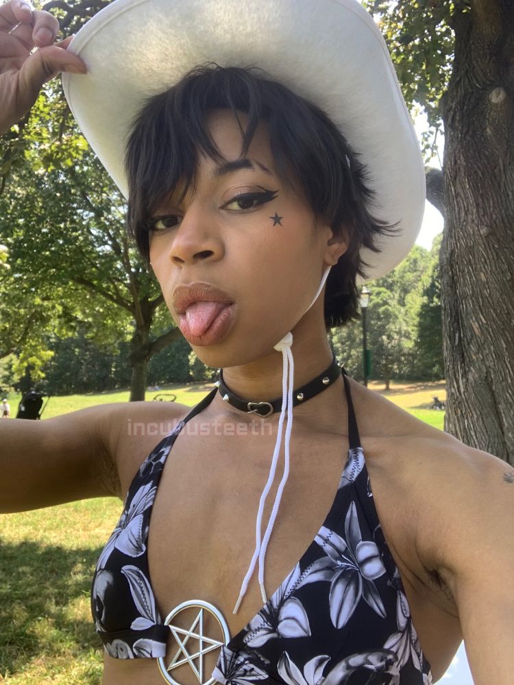 Danny tips the brim of his white cowboy hat while cutely sticking his tongue out. He’s standing in a sunny green park, surrounded by trees and grass. Danny is wearing sharp eyeliner, a small studded choker, and a black bikini patterned with white lilies and skulls.