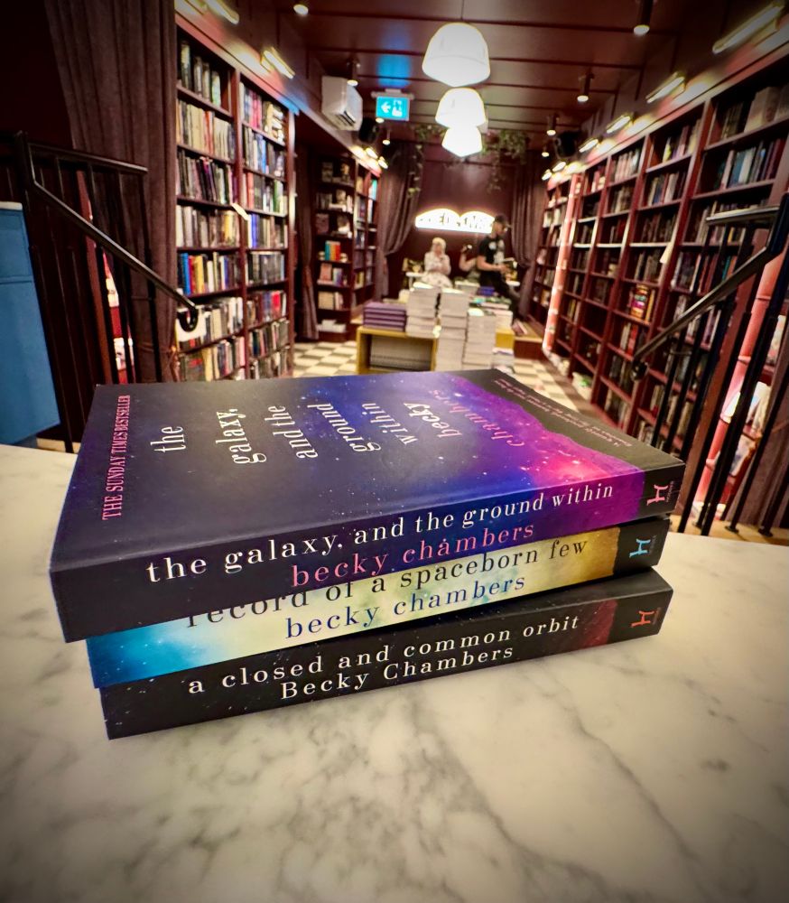 A photo of three novels by author Becky Chambers sitting on a cafe table in a bookstore, with well stocked bookshelves visible in the background.