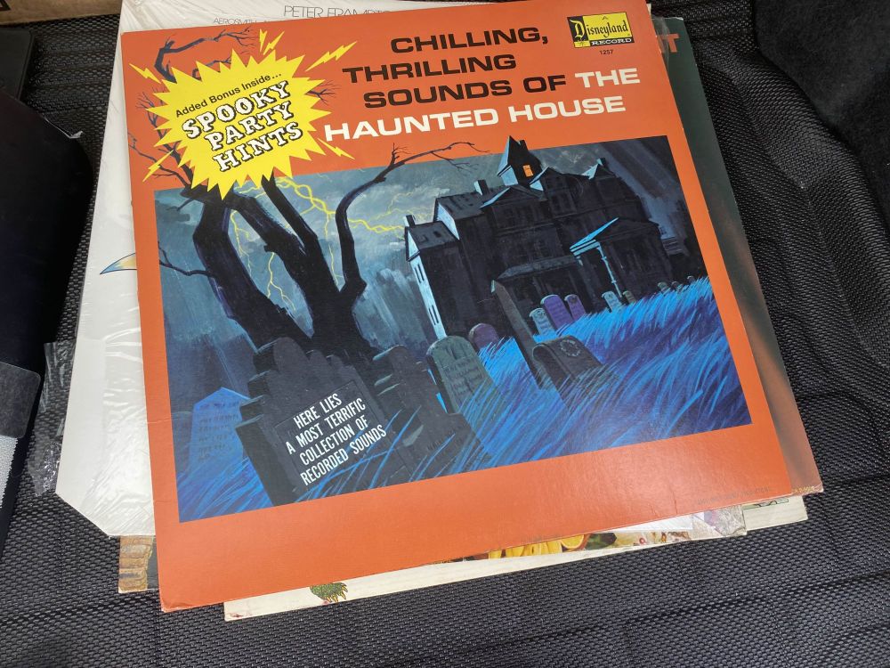 A photo of a vinyl album cover, with an orange background with the text "chilling, thrilling sounds of the haunted house" in the top right. A yellow shape is superimposed in the top left with the text "Added bonus inside, spooky party hints". below is a drawing of a graveyard with a tree and a mansion behind it, along with lightning. one one of the graves is the text "here lies a most terrific collection of recorded sounds".