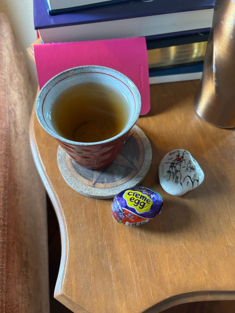A small ceramic cup of light-colored tea on a coaster next to a wrapped Cadbury creme egg and a rock painted with a line drawing of the monasteries of Meteora (Greece) with a pile of books and a metal water cup in the background, all on a little, wooden side table.