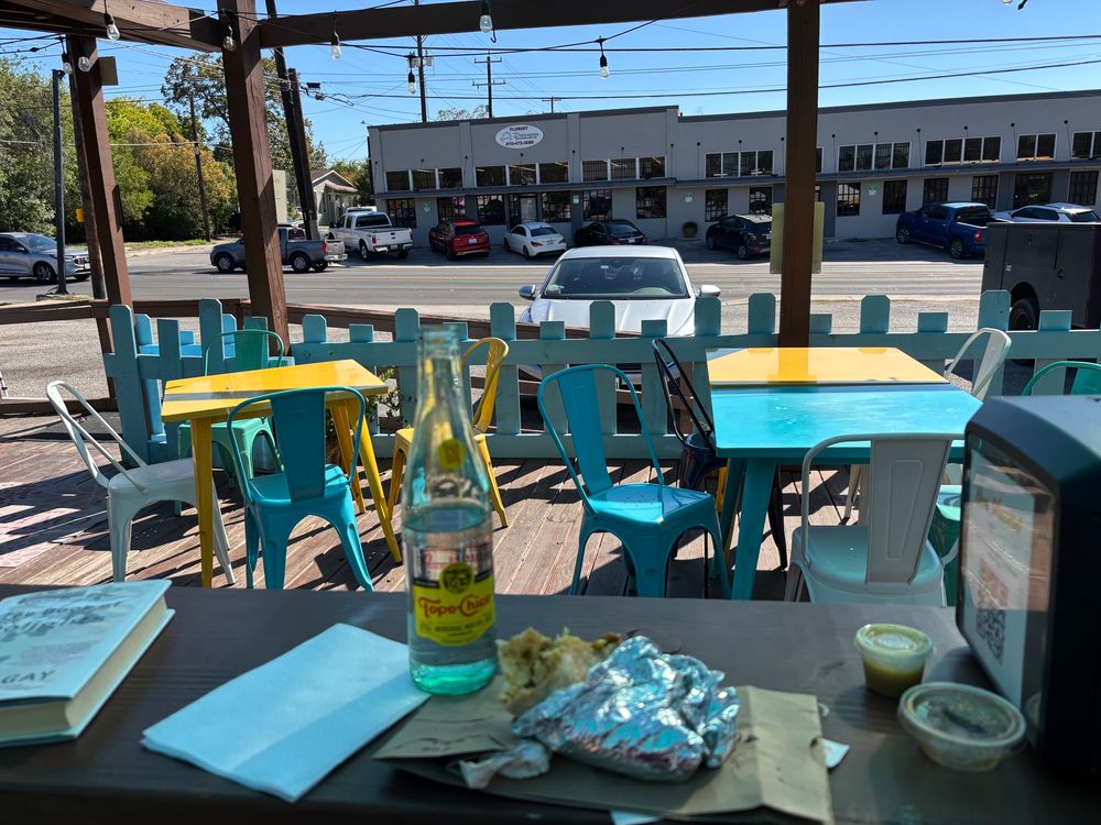 Outdoor wooden porch with colorful tables and chairs, facing the street with cars parked on both sides. On the table in front of me is a Carmen taco, a Topo Chico, some salsa, and a copy of Ross Gay’s “The Book of Delights,” which this certainly qualifies as. ❤️❤️