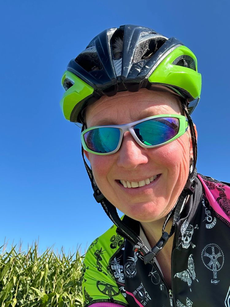 Erika wearing a green bike helmet, bike jersey, and sunglasses, posing in front of a mature cornfield.
