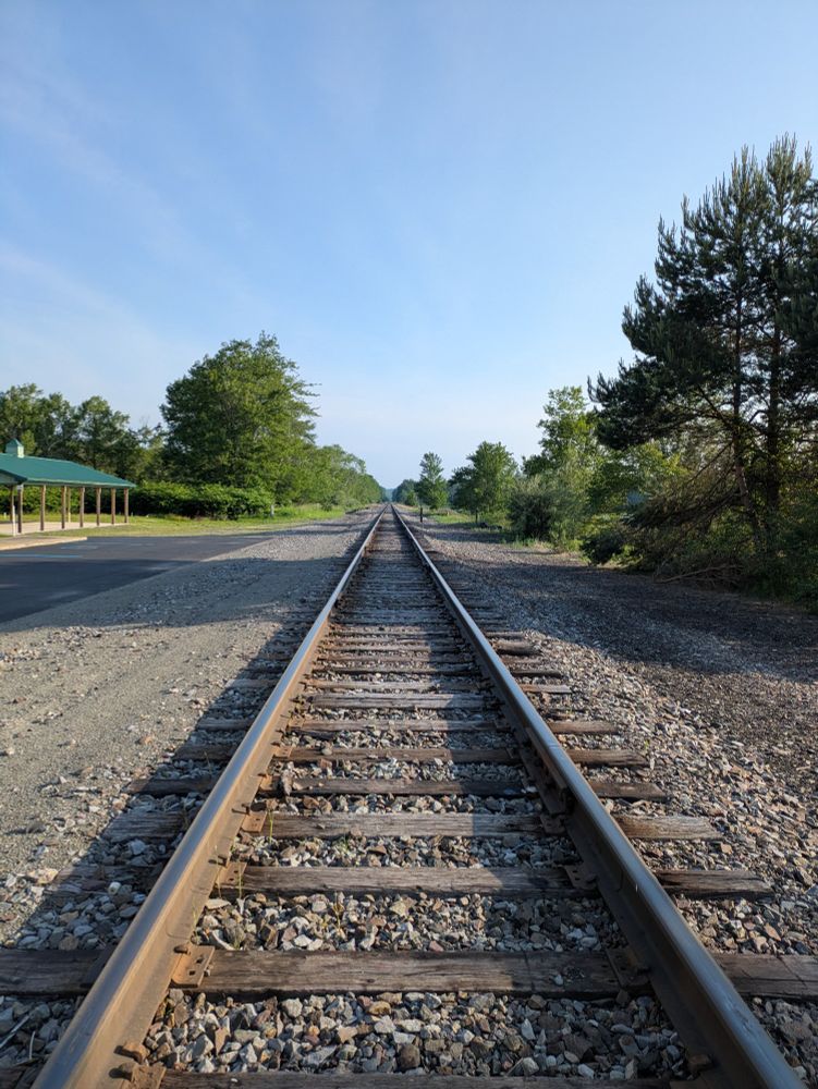 A long railroad track extends to the horizon, surrounded by trees, a small parking lot and a pavilion. 