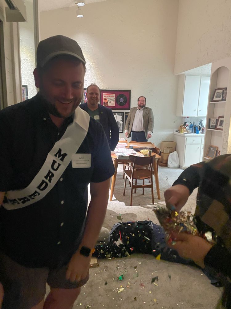 A smiling man surrounded by confetti wearing a sash that says “murderer” with a stunt dummy on the floor behind him.