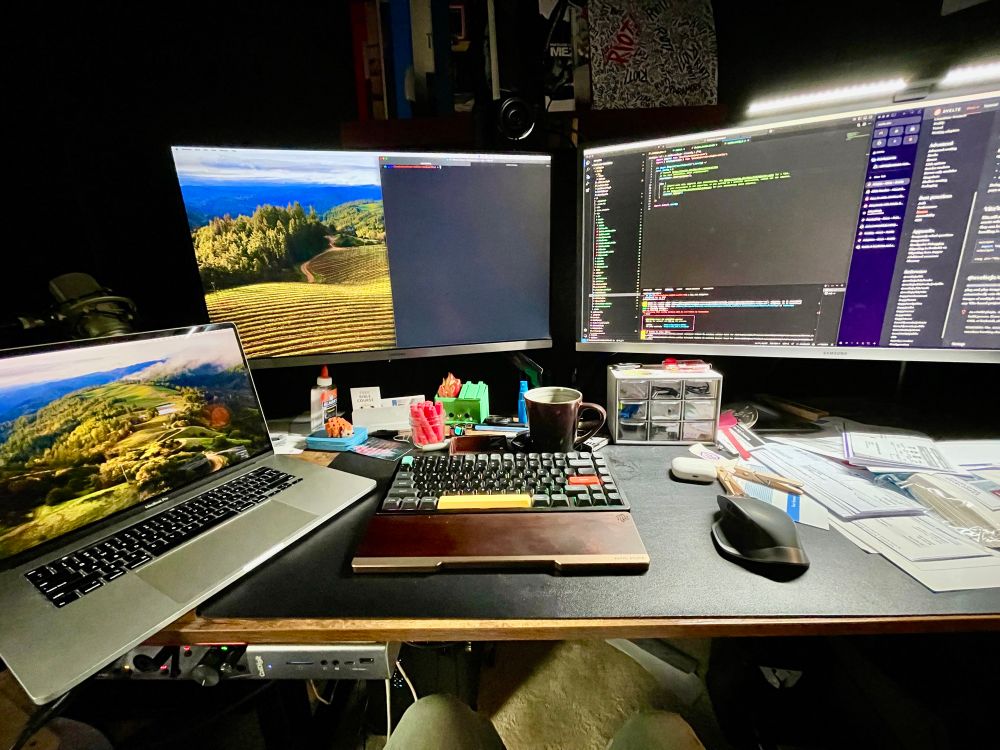 It’s my desk and it’s a mess. Two monitors and a laptop. A bar light over one of them. A coffee mug and a mess of bills and other mail to the right. Somehow there’s a bottle of Elmer’s glue too?