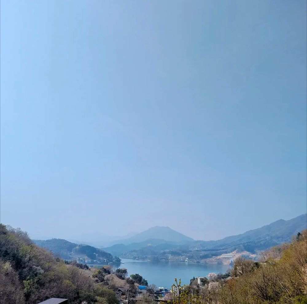 A photo of a lake surrounded by hills. The sky is a clear blue and the hills fade off into the distance. It is spring, and the hills are covered in green trees and flowering cherry blossoms.